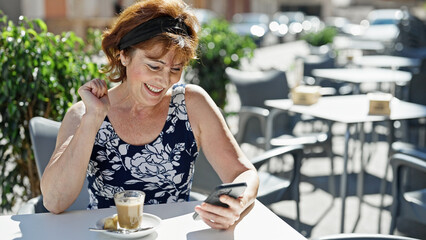 Middle age woman using smartphone with winner gesture at coffee shop terrace