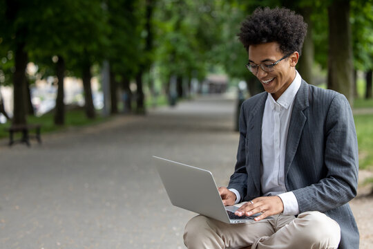 Curly-haired smiling young man working on laptop in the park - Powered by Adobe