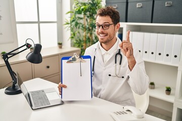 Young hispanic man wearing doctor stethoscope holding clipboard surprised with an idea or question pointing finger with happy face, number one