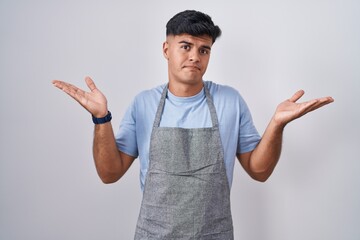 Hispanic young man wearing apron over white background clueless and confused expression with arms...