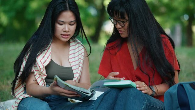 Two confident asian female students reading book while sitting on the grass at picnic