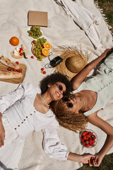 top view of pleased african american women laying on blanket near straw hat and food, summer picnic