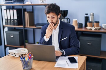 Handsome latin man working at the office using laptop covering mouth with hand, shocked and afraid...