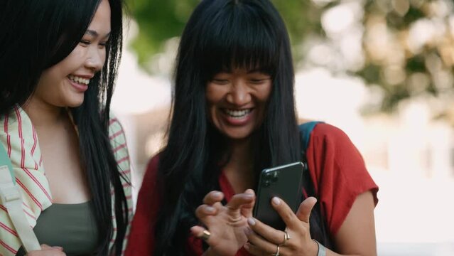 Two laughing asian women looking at photos on cell phone outdoors at summertime 