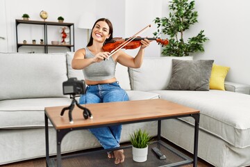 Young beautiful hispanic woman recording violin class sitting on sofa at home