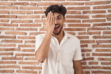 Arab man with beard standing over bricks wall background covering one eye with hand, confident smile on face and surprise emotion.