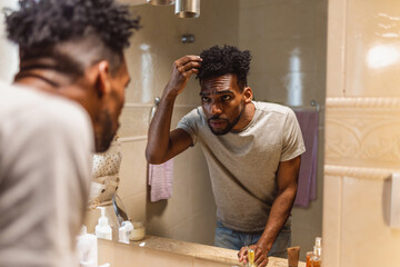 Homem negro jovem arrumando o cabelo no espelho do banheiro. Visão sobre o ombro.