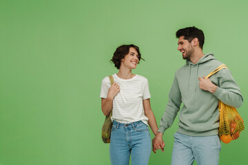 Smiling man and woman with mesh shopping bags holding hands while standing isolated over green wall