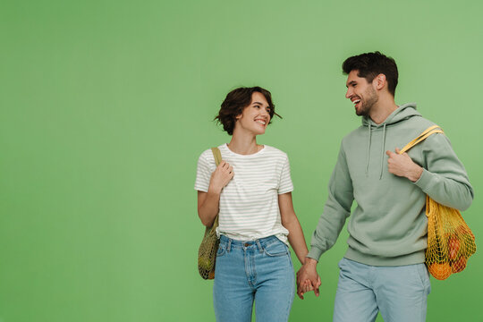Smiling man and woman with mesh shopping bags holding hands while standing isolated over green wall