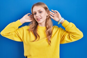 Young caucasian woman standing over blue background smiling pulling ears with fingers, funny...