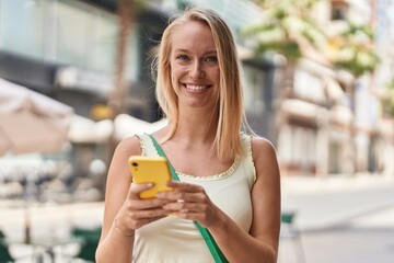 Young blonde woman smiling confident using smartphone at street