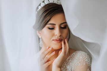 Female beauty. Cute woman at home. The bride poses under a veil. A stylish woman wears a white dress. Professional make-up and hair. On the bride's head is a crown and a long veil.