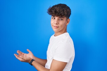 Hispanic teenager standing over blue background pointing aside with hands open palms showing copy...