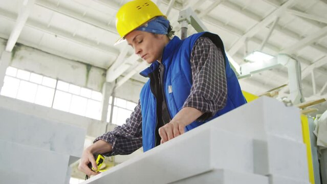 Low angle view of woman in hardhat and workwear using measuring tape to learn length and width of marble slab at work in stone factory