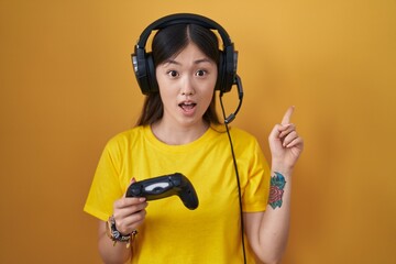 Chinese young woman playing video game holding controller surprised pointing with finger to the side, open mouth amazed expression.