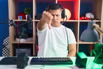 Young hispanic man playing video games looking unhappy and angry showing rejection and negative with thumbs down gesture. bad expression.