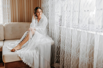 Fototapeta premium A stunning young bride poses by the window holding her veil in her hands. A beautiful woman of Caucasian appearance in an elegant white robe and luxurious hairstyle. Wedding makeup