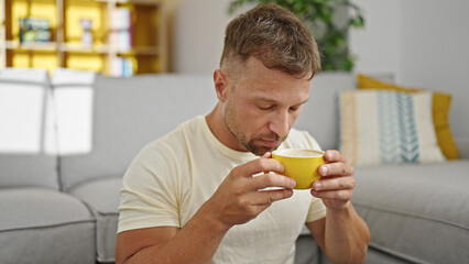 Young man smelling cup of coffee sitting on floor at home