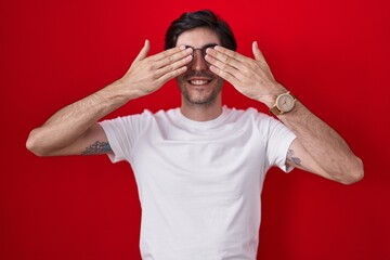 Young hispanic man standing over red background covering eyes with hands smiling cheerful and...