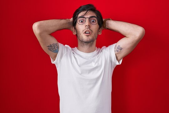 Young hispanic man standing over red background crazy and scared with hands on head, afraid and surprised of shock with open mouth