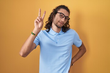 Young hispanic man standing over yellow background smiling looking to the camera showing fingers...