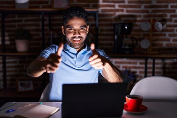 Hispanic man with beard using laptop at night approving doing positive gesture with hand, thumbs up smiling and happy for success. winner gesture.