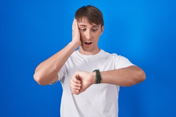 Caucasian blond man standing over blue background looking at the watch time worried, afraid of getting late