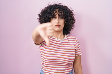 Young middle east woman standing over pink background looking unhappy and angry showing rejection and negative with thumbs down gesture. bad expression.