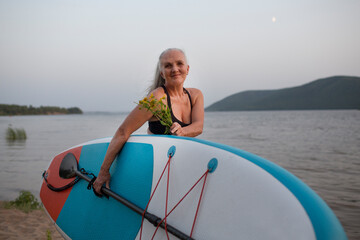 a smiling senior woman with gray hair holding sup board on the seashore at sunset. copy space. Slow life. Enjoying the little things. spends time in nature in summer