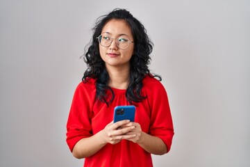 Young asian woman using smartphone typing message smiling looking to the side and staring away thinking.