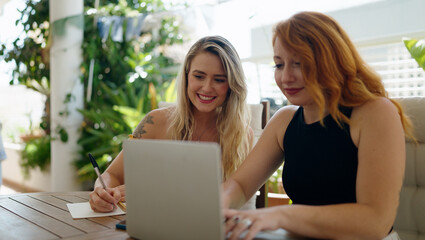 Two women using laptop writing on notebook sitting on table at home terrace