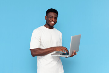 african american guy in white t-shirt uses laptop and smiles on blue isolated background, man...