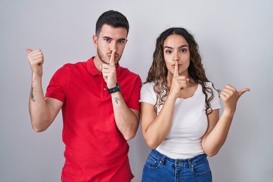Young hispanic couple standing over isolated background asking to be quiet with finger on lips pointing with hand to the side. silence and secret concept.