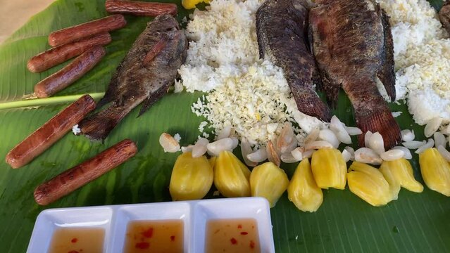 Fried fish and grilled sausages are laid out on rice, which is on a banana leaf with fruits. tropical food. High quality FullHD footage