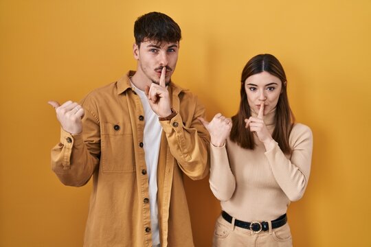 Young hispanic couple standing over yellow background asking to be quiet with finger on lips pointing with hand to the side. silence and secret concept.