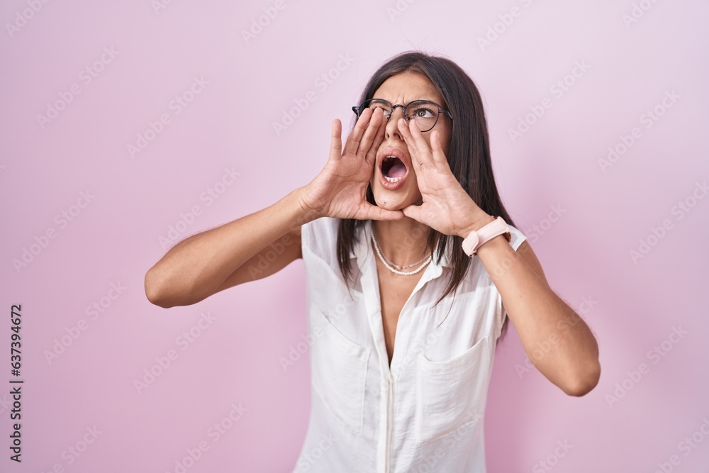 Poster Brunette young woman standing over pink background wearing glasses shouting angry out loud with hands over mouth