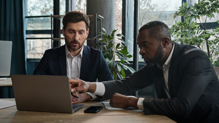Two diverse multiracial businessmen discuss online data project with laptop in office writing notes...
