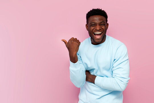 african american man in blue sweater laughs and points his hand to the side on pink isolated background