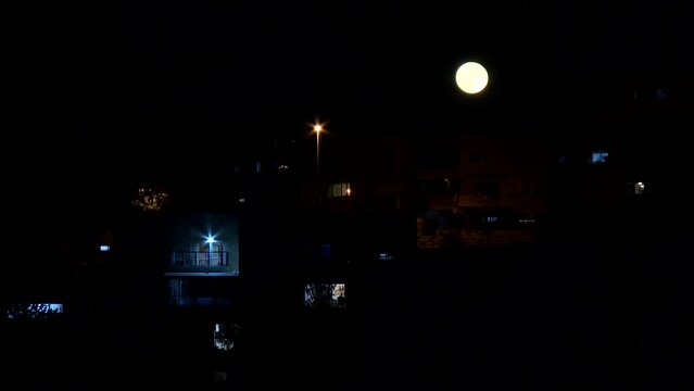 A dark scene, with few slightly lit balconies, and a full moon above them. an evening has fallen on the neighborhood. Jerusalem, Israel.