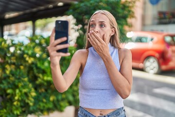 Young caucasian woman doing video call with smartphone covering mouth with hand, shocked and afraid...
