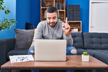 Hispanic man using laptop working from home surprised with an idea or question pointing finger with happy face, number one