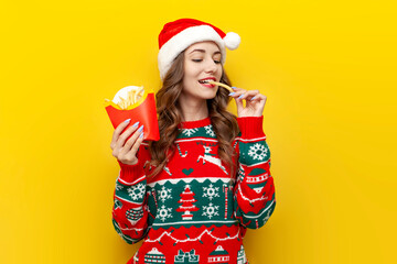 young girl in christmas sweater and santa claus hat eats fried french fries on yellow isolated...