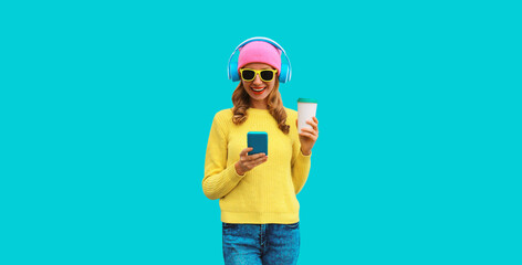 Portrait of stylish modern happy young woman listening to music in headphones with smartphone and cup of coffee wearing colorful pink hat and yellow sweater on blue background