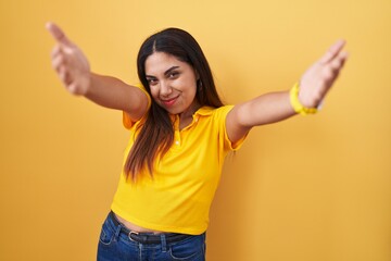 Young arab woman standing over yellow background looking at the camera smiling with open arms for hug. cheerful expression embracing happiness.