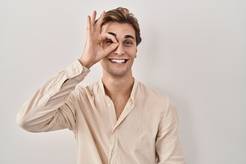 Young man standing over isolated background doing ok gesture with hand smiling, eye looking through fingers with happy face.