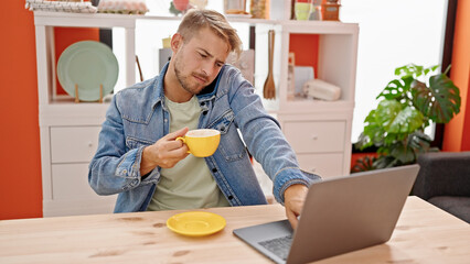 Young caucasian man drinking coffee talking on smartphone using laptop at dinning room