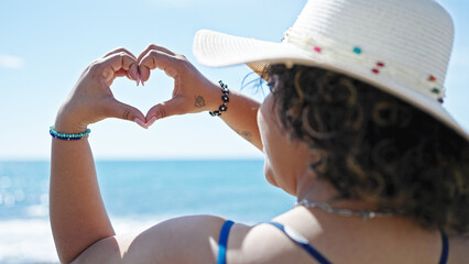 Young beautiful latin woman tourist wearing summer hat doing heart symbol with hands at seaside