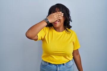 Hispanic woman standing over blue background smiling and laughing with hand on face covering eyes...