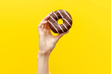hand raises and holds one sweet donut in chocolate icing on yellow isolated background