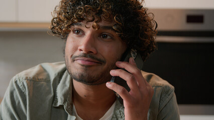 Cheerful Indian young man with curly hair talking phone in home kitchen. Happy friendly Hispanic guy talk with friends by smartphone Arabian male speaking smiling laughing pleasant mobile conversation